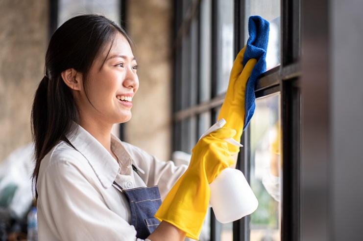 A woman wearing yellow rubber gloves, representing a janitorial service, cleans a window with a blue cloth and a spray bottle, smiling slightly.