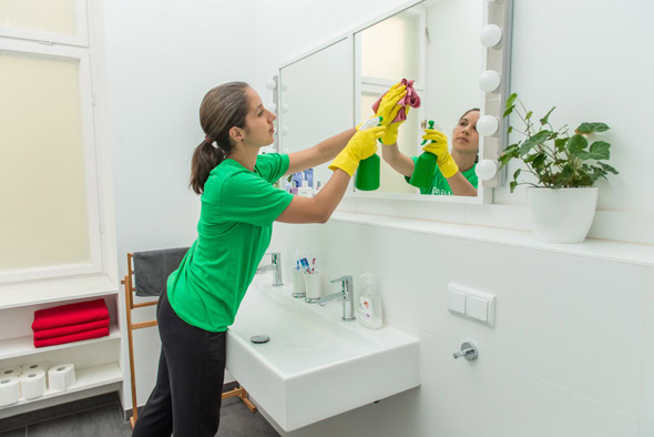 A woman wearing yellow gloves cleans a bathroom mirror with a cloth and spray bottle in a modern, white bathroom, demonstrating professional janitorial service.